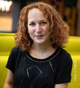 Sarah is a white person with auburn, curly hair. She’s sitting on a yellow sofa, wearing a black top, and smiling at the camera. Photo credit: Tony C French